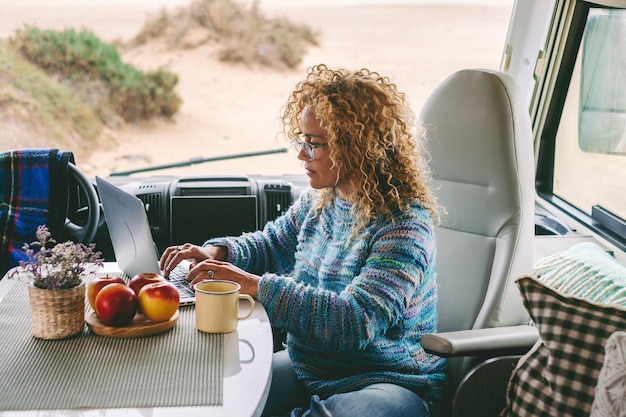 Une touriste moderne travaillant sur un ordinateur portable assis à l'intérieur d'un camping-car moderne avec vue sur la plage parking en arrière-plan