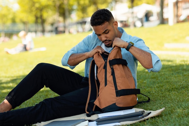 Un touriste noir cherche et sort quelque chose de son sac à dos assis dans un parc ensoleillé à l'extérieur