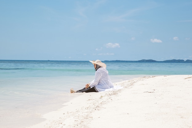 Touriste portant un chapeau d'été profitant du paysage marin et assis sur une plage de sable blanc en été