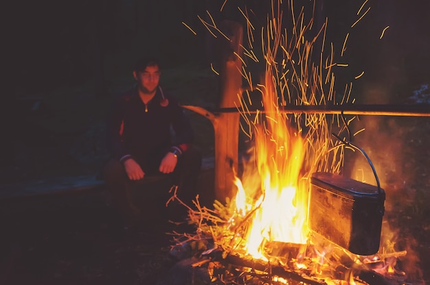 Le touriste de randonnée se repose dans son camp la nuit près du feu de camp. Guy boit du thé au coin du feu. Un homme regarde un feu de nuit.