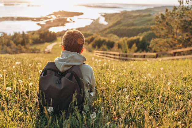 Touriste avec sac à dos assis au sommet d'une colline dans un champ d'herbe et profitant d'une belle vue sur le paysage Vue arrière d'un randonneur adolescent se reposant dans la nature Mode de vie actif Concept de voyage local