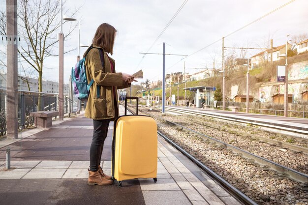 Une touriste avec un sac à dos et une grosse valise jaune se tient sur la plate-forme et attend le train