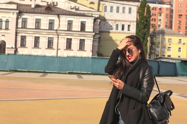 Une touriste souriante de femme brune écoute de la musique sur un téléphone portable, debout dans la rue avec un sac à dos