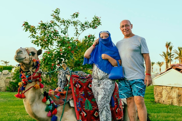 Touristes de couple de famille à côté de la femme de chameau couchée décorée avec le visage fermé de cap.