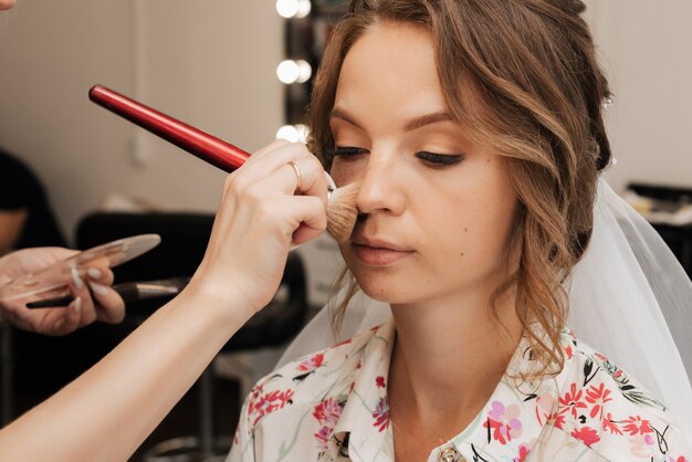 Tournage dans un salon de beauté. maquilleuse applique le maquillage à une jeune mariée belle fille.