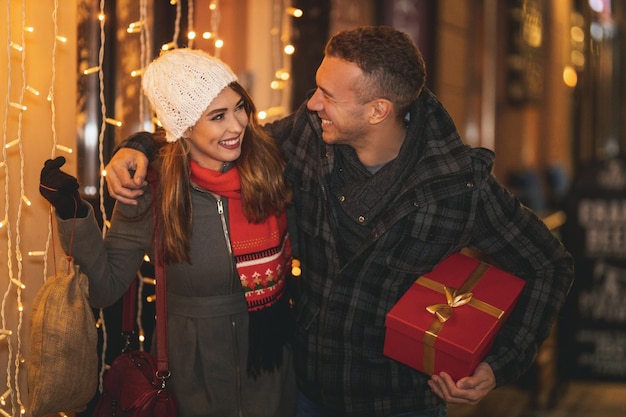 Le tournage d'un jeune couple joyeux s'amuse dans la rue de la ville la nuit de Noël. Ils rient et achètent des cadeaux pour les vacances à venir.