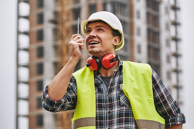 Tourner à gauche jeune constructeur gai dans un casque de protection jaune uniforme de travail parlant au grutier