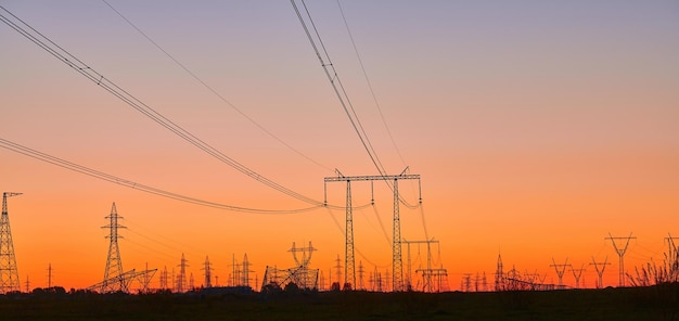 Tours électriques à haute tension sur le terrain au coucher du soleil et au ciel bleu Vue panoramique des silhouettes sombres des lignes électriques au lever du soleil orange Réseau de transmission et de distribution de production d'électricité