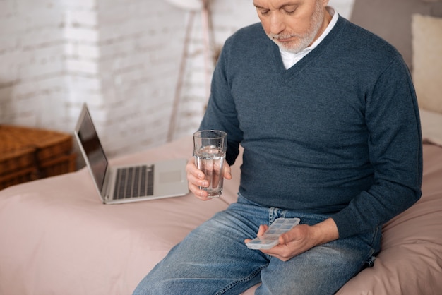 Tous les jours. Portrait of senior man holding case avec oreiller et verre d'eau alors qu'il était assis sur le lit.