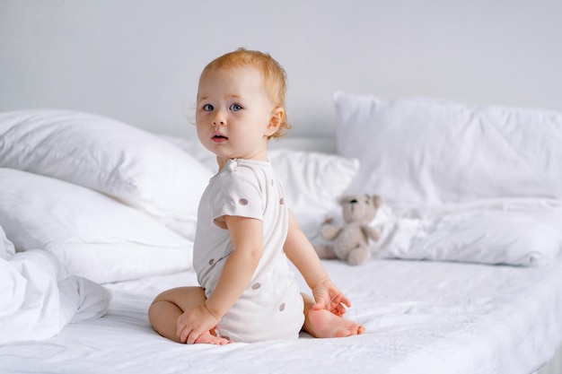 Tout-petit blond ou blond assis avec son dos sur un lit blanc dans une chambre lumineuse regardant la caméra