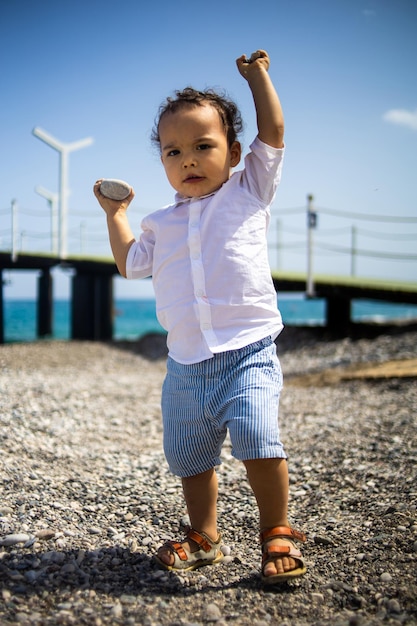 Un tout-petit garçon bouclé joue sur la plage de galets près de la mer bleue