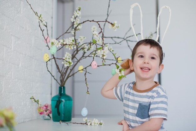 Un tout-petit garçon caucasien avec des oreilles de lapin drôles se tient près de l'arbre de pâques avec des oeufs colorés sur fond blanc