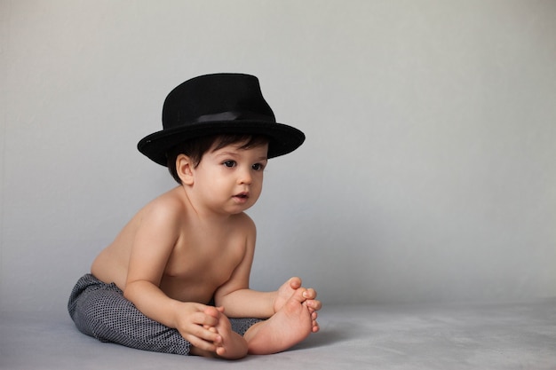 Tout-petit garçon en chapeau noir et pantalon noir avec un torse nu est assis sur un fond gris neutre.