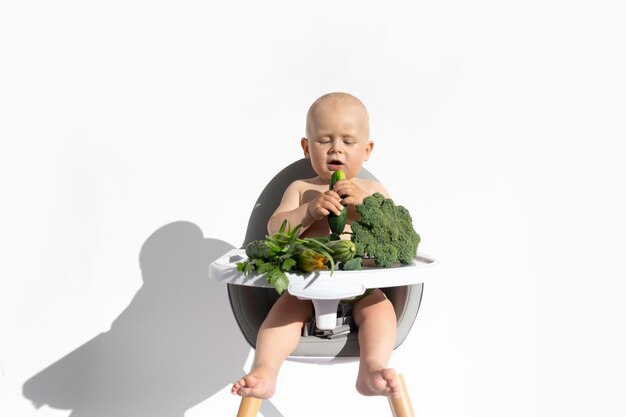 Tout-petit garçon dans la chaise d'alimentation mange des légumes biologiques salade concombres oignon brocoli ombre dure isolé sur fond blanc alimentation saine végétalienne alimentation concept d'aliments solides espace de copie