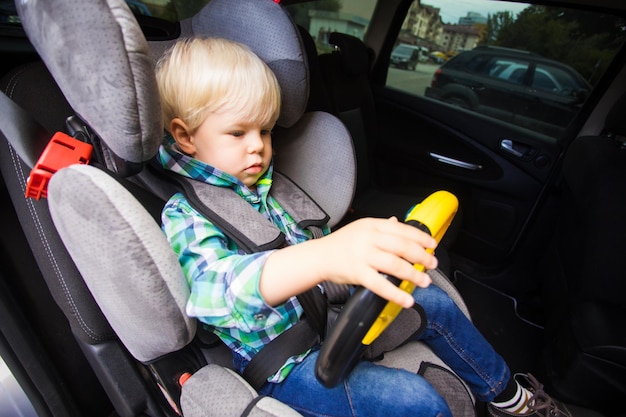 Le tout-petit garçon est assis attaché dans un siège de voiture et joue avec un volant de jouet