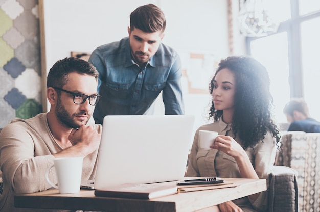 Tout revérifier. Jeune bel homme utilisant un ordinateur portable assis à la table du bureau lors d'une réunion d'affaires avec ses collègues