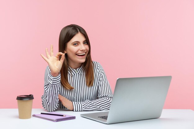 Tout va bien. Femme d'affaires positive et satisfaite en chemise rayée montrant un geste correct, un signe de main d'approbation, travaillant sur un ordinateur portable au bureau à domicile. Studio intérieur tourné isolé sur fond rose