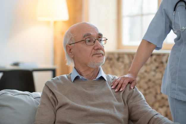 Tout va bien. Homme senior mélancolique bouleversé assis tout en regardant de côté et médecin mettant sa main sur son épaule