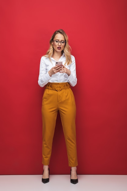 Sur toute la longueur d'une belle jeune femme blonde choquée debout isolé sur fond rouge, tenant un téléphone mobile