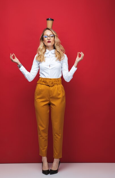 Sur toute la longueur d'une belle jeune femme blonde debout isolé sur fond rouge, équilibrage, tenant une tasse de café