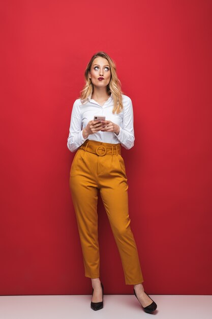 Sur toute la longueur d'une belle jeune femme blonde pensive debout isolé sur fond rouge, tenant un téléphone mobile