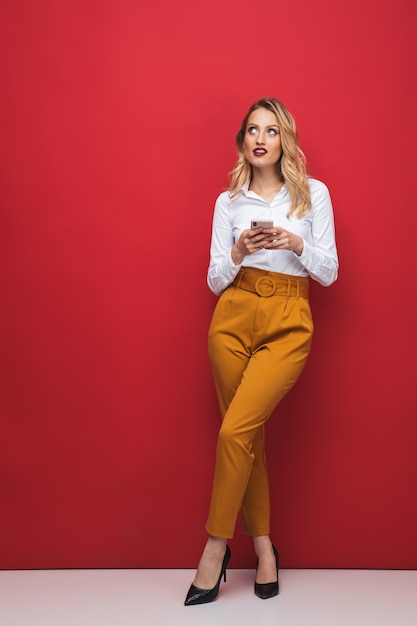 Sur toute la longueur d'une belle jeune femme blonde pensive debout isolé sur fond rouge, tenant un téléphone mobile