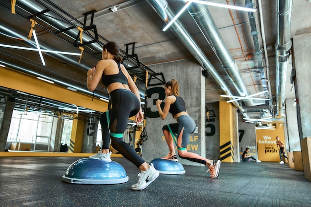 Toute la longueur de deux jeunes femmes athlétiques en vêtements de sport faisant de l'exercice avec des bandes de résistance à la gym crossfit, faisant des squats et des jambes d'entraînement. Entraînement, sport, bien-être et mode de vie sain