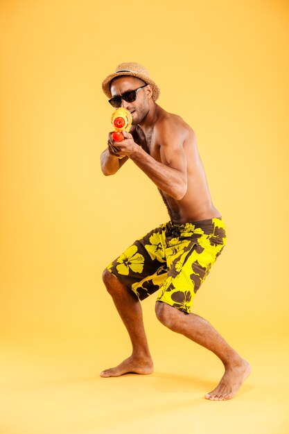 Sur toute la longueur de drôle jeune homme afro en maillot de bain tir avec pistolet à eau sur mur orange