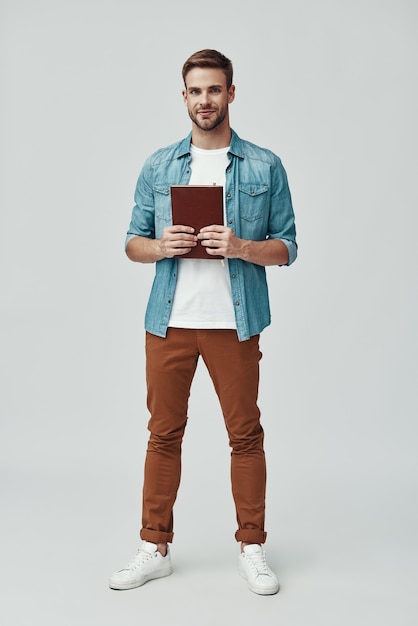 Toute la longueur du beau jeune homme regardant la caméra et souriant en se tenant debout sur fond gris