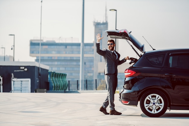 Sur toute la longueur du bel homme d'affaires barbu caucasien avec des lunettes de soleil prenant son sac en cuir du coffre et en agitant.