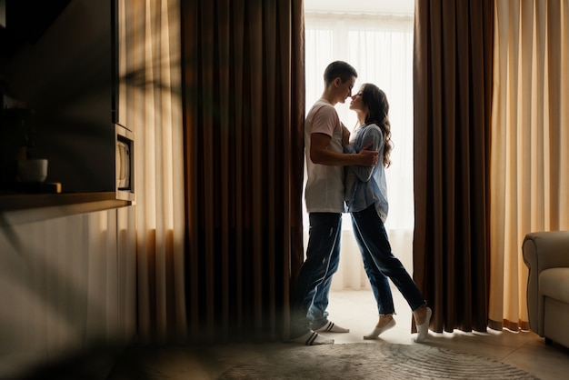 Toute la longueur du couple d'amoureux s'embrassant dans la cuisine à la maison tout en se regardant contre la fenêtre. Relation, concept d'amour. Discret.