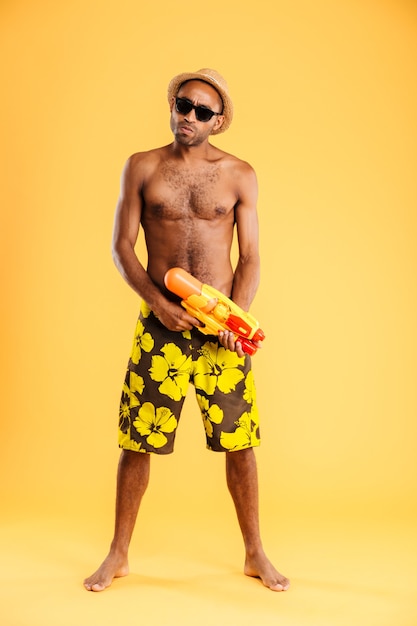 Sur toute la longueur du jeune homme afro drôle en maillot de bain tenant un pistolet à eau sur un mur orange