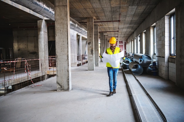 Toute La Longueur Du Travailleur De La Construction En Vêtements De Travail à Marcher Dans Le Bâtiment Dans Le Processus De Construction Et à La Recherche De Plans.