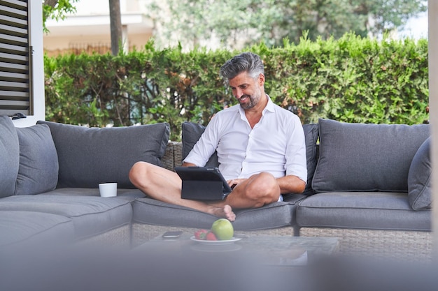 Toute la longueur d'un entrepreneur masculin positif naviguant sur une tablette et souriant tout en travaillant sur un projet à distance dans une cour clôturée par une haie