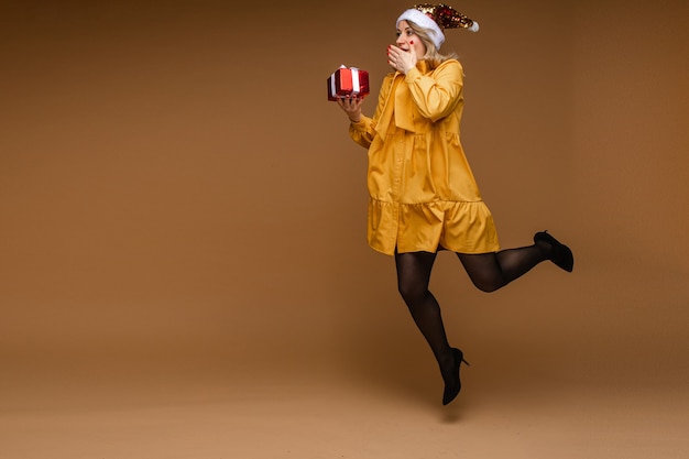 Toute la longueur de la femme blonde de race blanche en robe jaune, collants, talons et bonnet de Noel étincelant transportant un cadeau de Noël enveloppé dans la bouche couvrant l'excitation