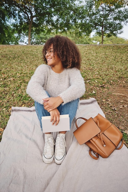 Toute la longueur d'une femme ethnique heureuse en tenue décontractée souriante et regardant ailleurs assise sur une couverture sur l'herbe avec un livre et embrassant les genoux dans la nature le week-end