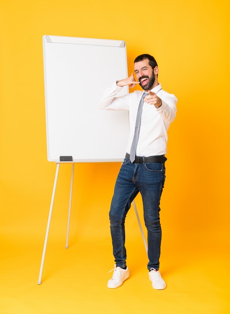 Toute la longueur de l'homme d'affaires donnant une présentation sur tableau blanc sur mur jaune isolé faisant un geste de téléphone et pointant le devant