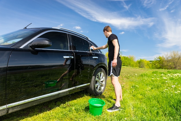 Toute la longueur de l'homme lave la voiture avec une éponge savonneuse dans les champs verts par une belle journée ensoleillée avec un ciel bleu
