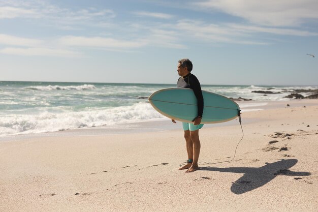 Toute la longueur d'un homme senior biracial transportant une planche de surf debout à la plage regardant loin contre le ciel