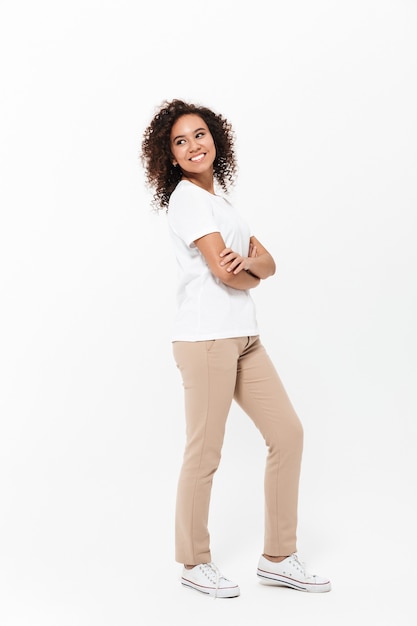 Toute la longueur d'une jeune femme africaine heureuse portant des vêtements décontractés isolés sur un mur blanc, les bras croisés