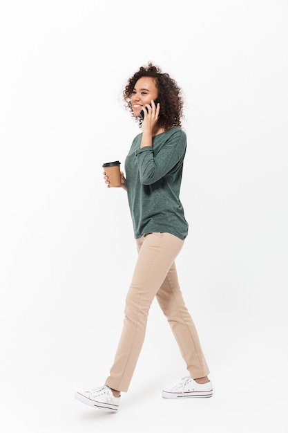 Toute la longueur d'une jeune femme africaine heureuse portant des vêtements décontractés isolés sur un mur blanc, parlant au téléphone portable, buvant du café à emporter