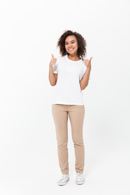 Toute la longueur d'une jeune femme africaine heureuse portant des vêtements décontractés isolés sur un mur blanc, les pouces vers le haut