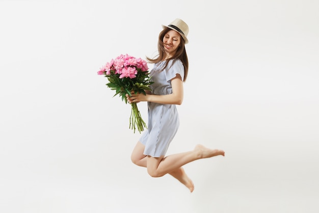 Toute la longueur d'une jeune femme heureuse et amusante en robe bleue, chapeau tenant un bouquet de fleurs de pivoines roses, sautant isolé sur fond blanc. Saint-Valentin, concept de vacances de la Journée internationale de la femme.