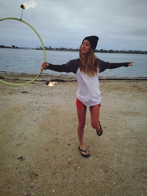 Toute la longueur d'une jeune femme avec un hula hoop de feu sur la plage contre le ciel