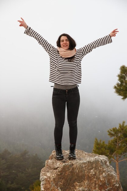 Photo toute la longueur d'une jeune femme insouciante avec les bras levés debout sur un rocher par temps brumeux