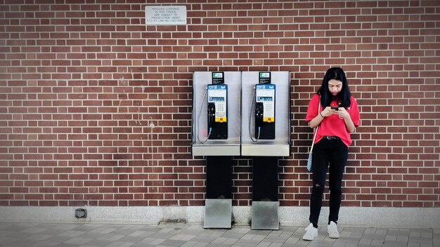 Photo toute la longueur d'une jeune femme utilisant le téléphone par cabine téléphonique contre un mur de briques