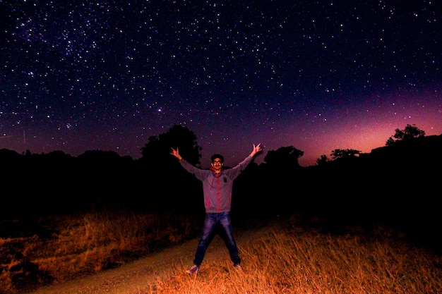 Photo toute la longueur d'un jeune homme avec les bras tendus debout sur le champ contre le champ d'étoiles la nuit