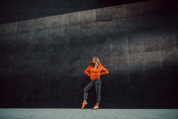 Toute la longueur de la jolie femme caucasienne fit Sportswear posant en se tenant debout contre le mur noir.