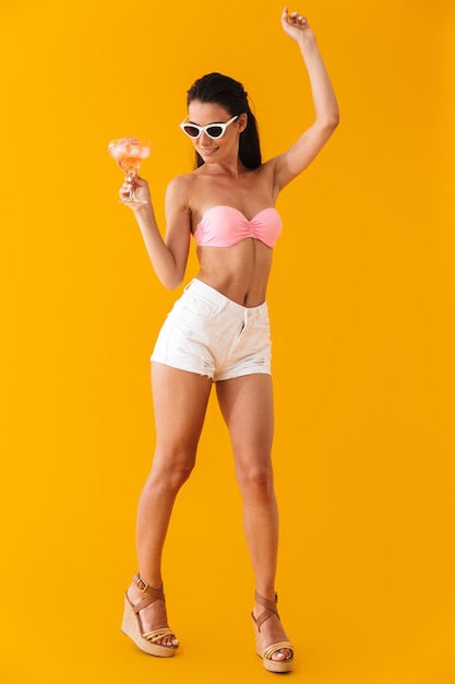 Toute la longueur d'une jolie jeune fille joyeuse portant un bikini debout isolé sur un mur jaune, tenant un cocktail en dansant