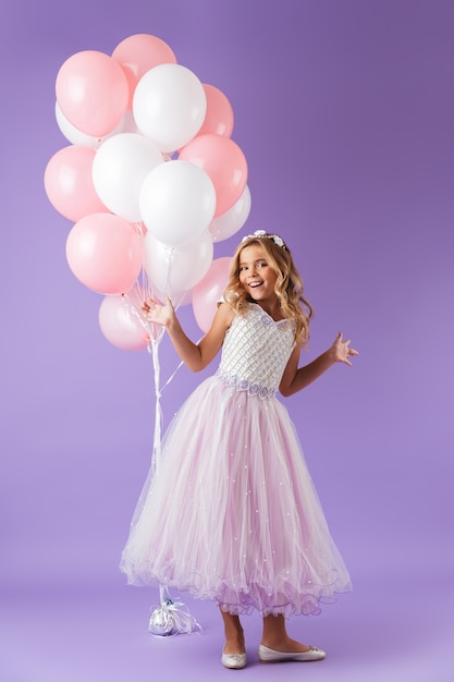 Sur toute la longueur d'une jolie petite fille habillée en robe de princesse debout isolé sur mur violet, tenant un bouquet de ballons